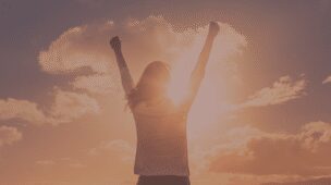 Mulher de braços abertos contra o sol com um céu azul e poucas nuvens no fundo